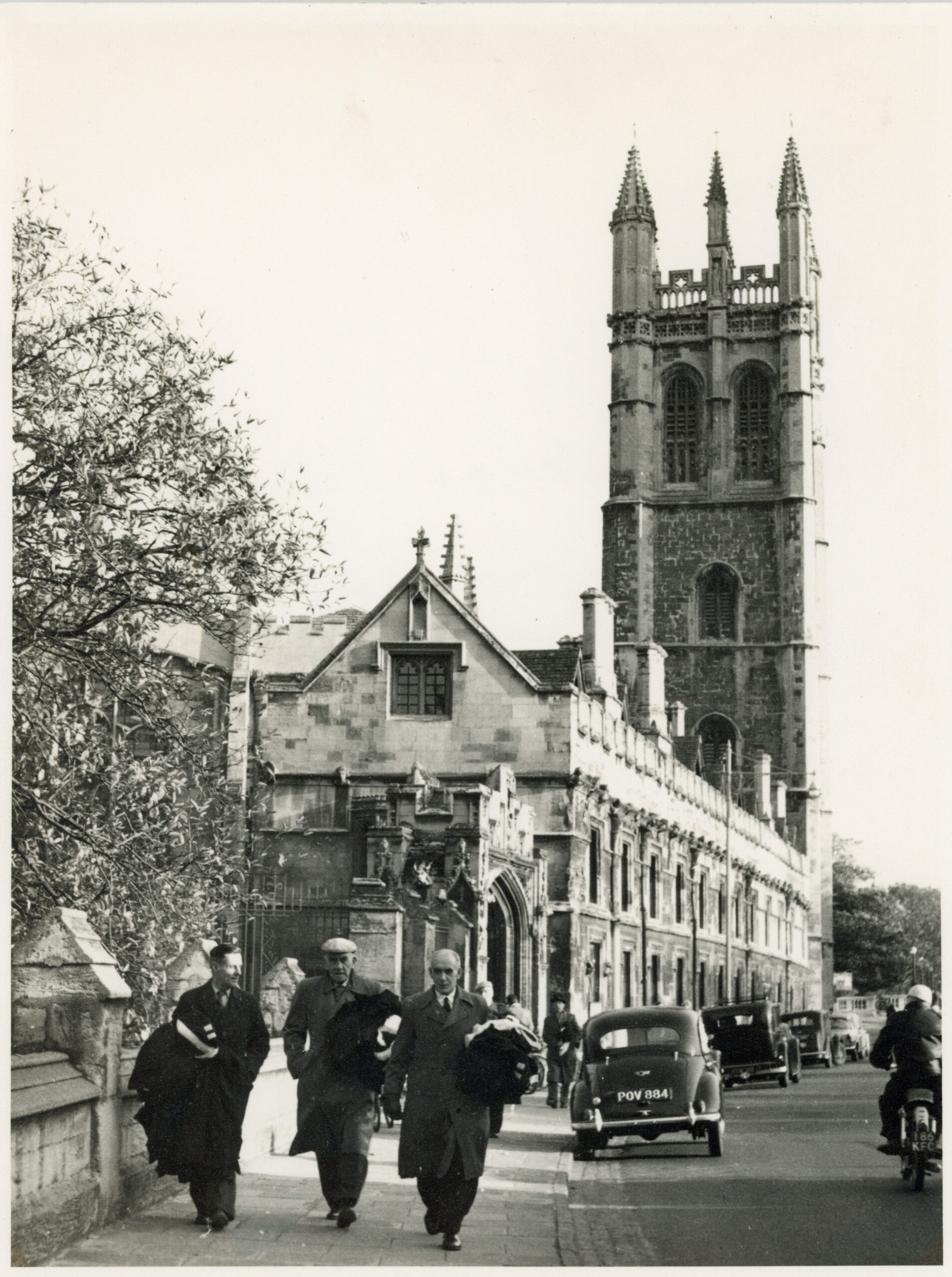Black-and-white photograph showing Reg Betnay, Magdalen College scout, carrying gowns to Fellows involved in a graduation ceremony in 1961. Betnay and his two companions are walking west along the High Street, Oxford, and Magdalen's Great Tower is seen in the background.