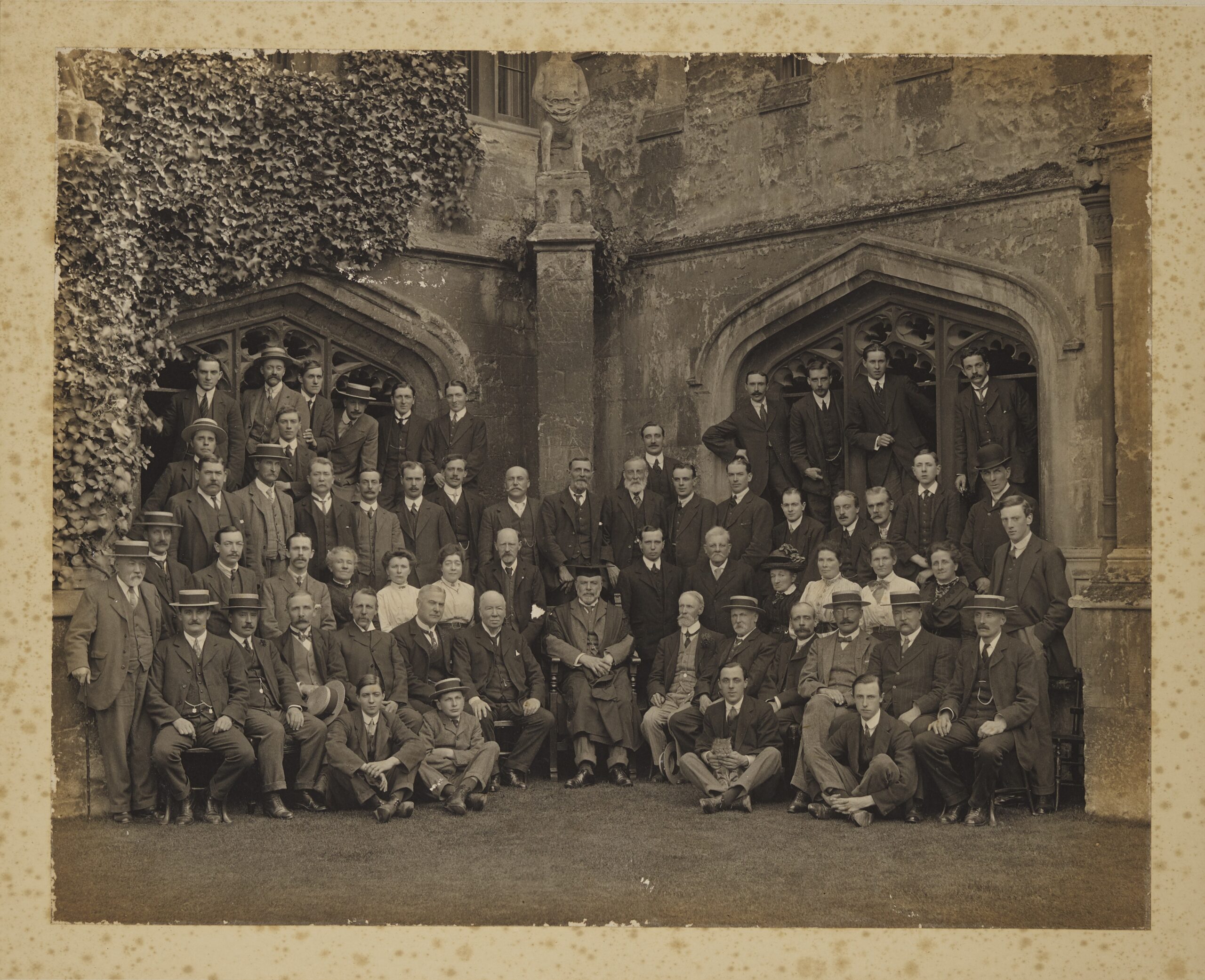 Group of photograph of Magdalen College servants in Cloisters with President Warren seated in the middle, circa 1910-1914. There are 60 sitters, including 7 women and a tabby cat. The photo is undated, and no names are given, but Richard Gunstone (“Gunner”), JCR Steward 1880-1914, is seated to Warren’s left.