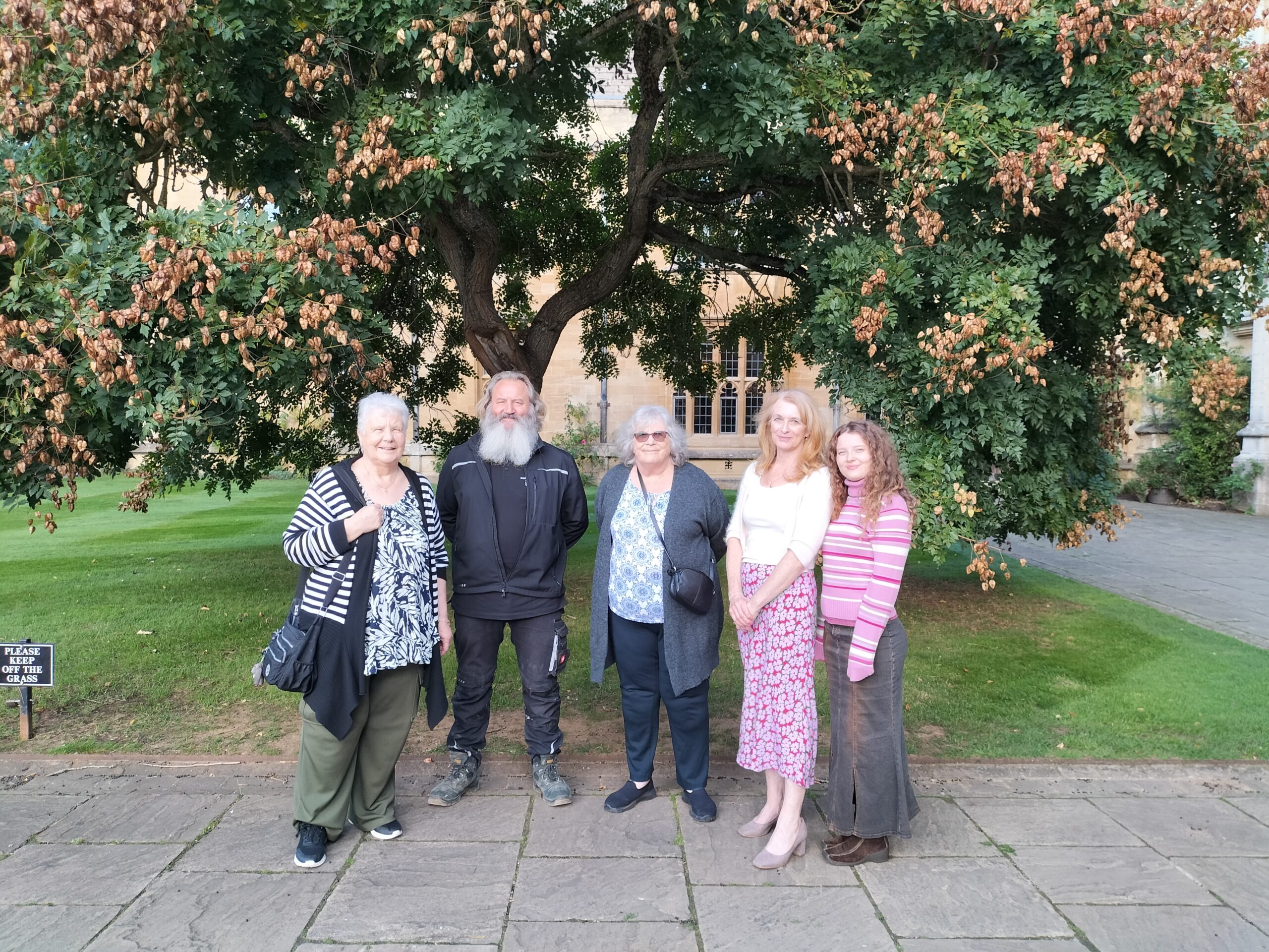 Colour photograph of Magdalen College Head Gardener David Craft, posed with three generations of the descendants of former Head Gardener Jack Sawyer, in front of the golden rain tree in St John's Quadrangle, 2025.